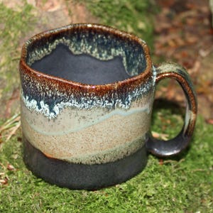 May include: A handmade ceramic mug with a square shape and a dark handle. The mug features a layered design with colours ranging from dark brown to tan, with a black base and interior. The mug is sitting on green moss.