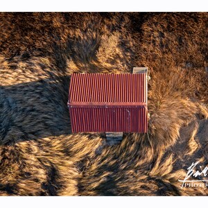May include: An aerial view of a small red metal-roofed building in a field of tall, dry grass. The grass is blowing in the wind, creating a swirling pattern around the building.