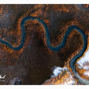 May include: Aerial view of a winding river flowing through a marsh. The water is dark blue, contrasting with the golden-brown reeds. The image is lit by the sun, creating a warm, inviting scene. The photographer's signature is in the bottom left corner.