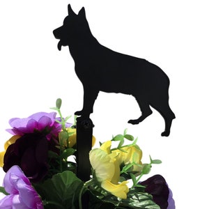 May include: A black metal silhouette of a German Shepherd dog stands atop a stake, designed for garden or flower bed decor. The dog is positioned above a colorful arrangement of purple, yellow, and green artificial flowers.