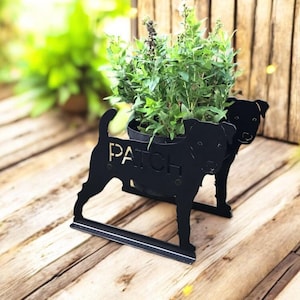 May include: Black metal dog-shaped planter with the word "PATCH" cut out. The planter holds a pot of green plants. The planter is on a wooden surface with a wooden fence in the background.