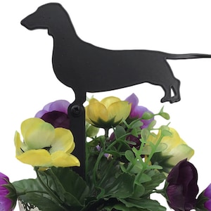 May include: A black metal dachshund silhouette garden stake. The dog is mounted on a vertical rod, and is placed in a pot of colorful artificial flowers. The flowers are yellow, purple, and green.