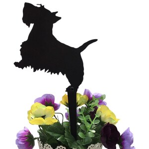 Pode incluir: Silhueta metálica preta de um cão Scottish Terrier, montada numa haste preta, sobre um arranjo floral. As flores são uma mistura de roxo, amarelo e verde, num recipiente decorativo de metal branco. O cão está numa pose brincalhona.