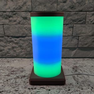 May include: A cylindrical lamp with a wooden base and a green, blue, and green gradient light. The lamp is on a light gray surface.