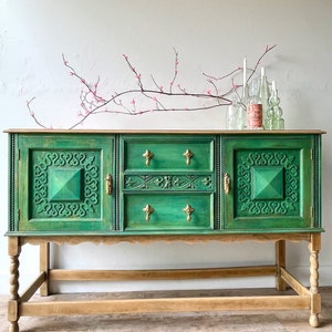 Könnte beinhalten: Ein vintage grünes Sideboard mit geschnitzten Details und Messingbeschlägen. Das Sideboard hat drei Schubladen und zwei Türen. Der Sockel ist aus Naturholz und hat gedrehte Beine.