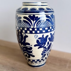 May include: A ceramic vase with a cream-coloured base and hand-painted deep blue floral motifs. The design features intricate botanical patterns, a checkered band around the middle, and a smaller checkered border near the base and rim.