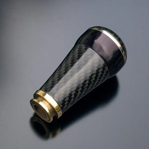 Pode incluir: Uma alavanca de câmbio de fibra de carbono preta com um anel dourado e uma moldura prateada.