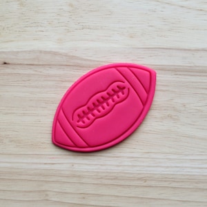 May include: A pink plastic cookie cutter shaped like a football. The football has a white seam down the middle.