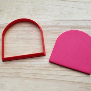 May include: A red plastic cookie cutter shaped like a rounded arch, and a pink fondant cutout in the same shape.