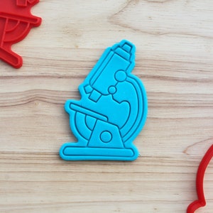 May include: A blue plastic cookie cutter shaped like a microscope. The cookie cutter is on a wooden surface.