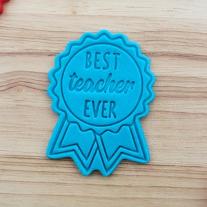 May include: A bright blue cookie cutter shaped like a circular award ribbon with scalloped edges and two tails at the bottom. The centre of the ribbon features the embossed text BEST teacher EVER in a combination of block and script lettering.