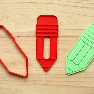 Op de afbeelding: Drie koekjesvormen in de vorm van een potlood, één rood, één groen en één rood met een holle kern. De vormen liggen op een houten oppervlak.