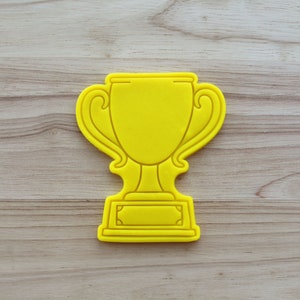 May include: Yellow plastic cookie cutter shaped like a trophy cup. The trophy has a large cup with two handles and a base with a rectangular space for a name.