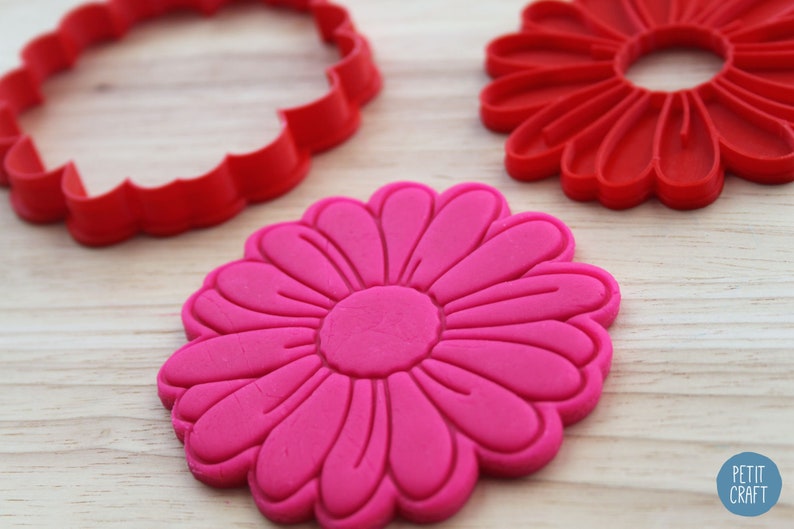 May include: Two red plastic flower-shaped cookie cutters and a pink flower-shaped cookie. The cookie cutters are on a wooden surface.