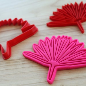 Puede incluir: Dos cortadores de galletas con forma de flor estilizada. Un cortador es rojo y el otro es rosa.