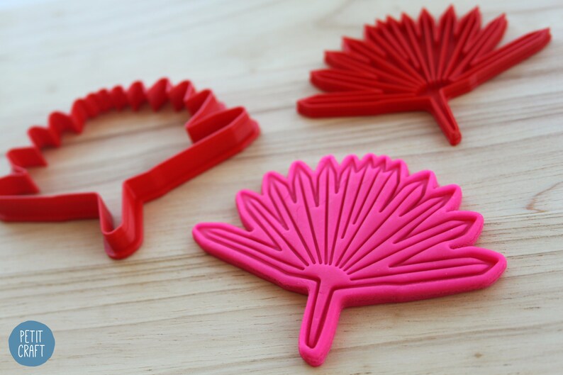 Puede incluir: Dos cortadores de galletas con forma de flor estilizada. Un cortador es rojo y el otro es rosa.