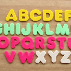 Puede incluir: Un conjunto de letras del alfabeto de colores hechas de plastilina. Las letras están ordenadas alfabéticamente y son de tres colores diferentes: amarillo, verde y rosa. Las letras están sobre una superficie de madera.