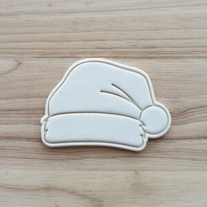 Op de afbeelding: Een witte koekjesvorm in de vorm van een kerstmuts met een gladde afwerking. Het ontwerp heeft een omgeslagen rand, een ronde pompon aan de zijkant en een gebogen bovendeel. Het object is gemaakt van stevig materiaal met scherpe randen.