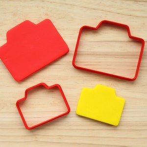 May include: Four red and yellow plastic cookie cutters shaped like cameras on a wooden surface. The cutters are different sizes.