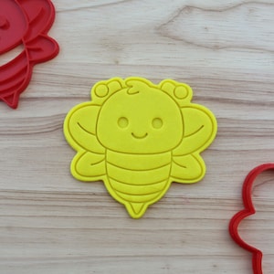 May include: A yellow bee-shaped cookie cutter with a smiling face and wing details. Two red bee-shaped cookie cutters are also visible. The cutters are on a light wood grain surface. The image is well-lit and suitable for baking-themed listings.