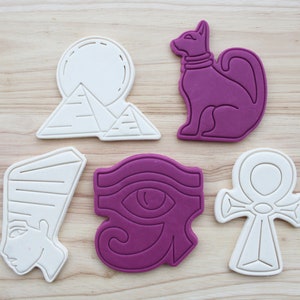 Op de afbeelding: Set van zes witte en paarse koekjesvormen met Egyptische thema's. De vormen omvatten een kat, een oog van Horus, een ankh, een piramide en een zon.