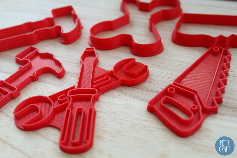 Może przedstawiać: Zestaw czerwonych plastikowych foremek do ciasteczek w kształcie r&oacute;żnych narzędzi, w tym młotka, klucza, śrubokręta i piły. Formy znajdują się na drewnianej powierzchni.