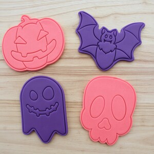 May include: Four plastic cookie cutters in Halloween shapes: a pink pumpkin, a purple bat, a purple ghost, and a pink skull. Each cutter features embossed details for facial features and wings. The top left corner includes a small blue circular logo with the text PETIT CRAFT.