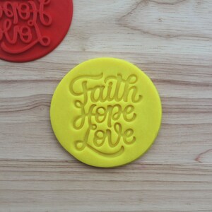 May include: Two circular cookie stamps with embossed lettering. One stamp is bright yellow and the other is red. Both feature the words Faith, Hope, and Love in a cursive script font. The stamps are designed for imprinting dough or clay.