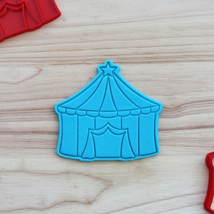 May include: A blue plastic cookie cutter shaped like a circus tent with a star on top. The tent has a flap in the front and a white stripe around the bottom.