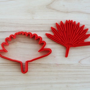 Puede incluir: Dos cortadores de galletas de pl&aacute;stico rojo, uno con forma de hoja de palmera de abanico y el otro con forma de flor redonda con tallo.