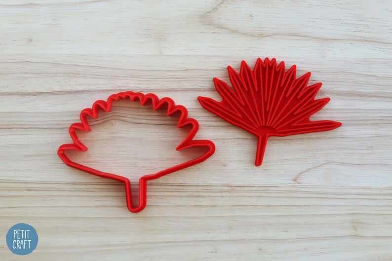 Puede incluir: Dos cortadores de galletas de pl&aacute;stico rojo, uno con forma de hoja de palmera de abanico y el otro con forma de flor redonda con tallo.
