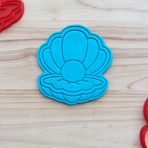May include: A blue plastic cookie cutter shaped like a seashell with a pearl inside. The cutter is on a light brown wooden surface.