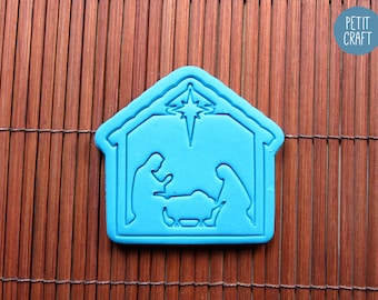 May include: A bright blue cookie cutter shaped like a nativity scene, featuring a stable outline, a star at the top, and silhouettes of two figures standing over a manger. The text PETIT CRAFT is visible in a blue circle in the upper right corner.