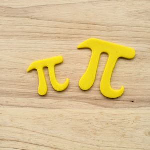 Könnte beinhalten: Zwei gelbe Tonmodelle des griechischen Buchstabens Pi, eines größer als das andere, auf einer hellbraunen Holzoberfläche.