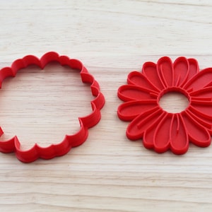 May include: Two red plastic cookie cutters, one shaped like a simple circle with scalloped edges and the other shaped like a daisy with a centre hole.