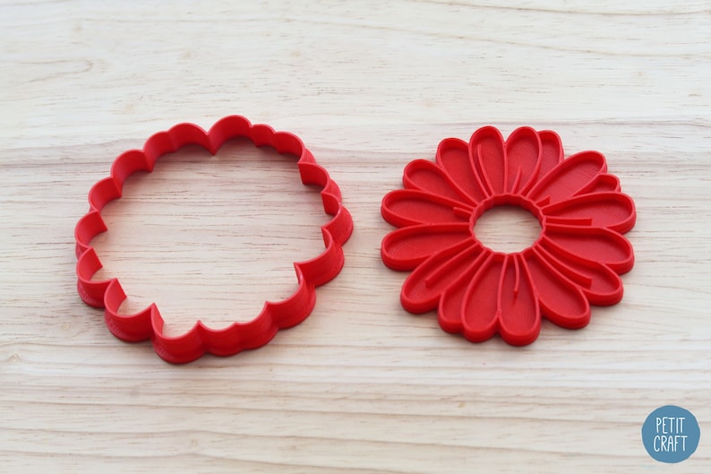 May include: Two red plastic cookie cutters, one shaped like a simple circle with scalloped edges and the other shaped like a daisy with a centre hole.