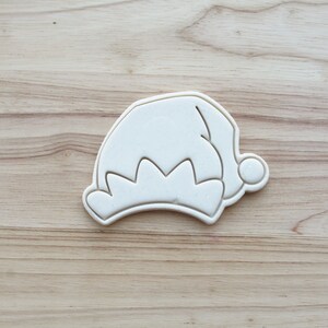 Op de afbeelding: Een witte plastic koekjesvorm in de vorm van een elfenmuts met een gekartelde rand en een pompon aan de punt. Het ontwerp heeft een duidelijke omtrek en reliëflijnen aan de binnenkant om de vorm van de muts te creëren. Het object is solide en ondoorzichtig.