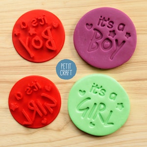 May include: Four round, colourful, silicone stamps with the text "it's a boy" and "it's a girl" in white. The stamps are on a wooden surface.