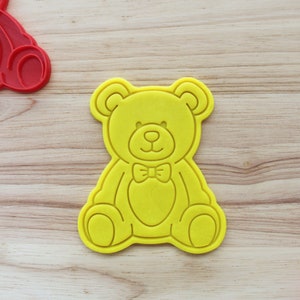May include: Yellow plastic cookie cutter shaped like a teddy bear with a bow tie. The bear is sitting with its legs crossed and has a simple, cartoon-like design.