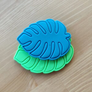 May include: Two leaf-shaped cookie cutters, one in bright blue and one in vibrant green, featuring detailed embossed veins and a distinct monstera leaf silhouette. The blue cutter is layered on top of the green cutter, both showing a smooth finish.