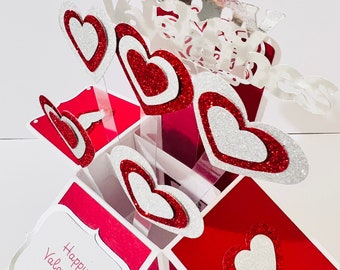 May include: A red and white pop-up card with the words "Happy Valentines Day" on the front. The card is decorated with glitter hearts and the words "Happy Valentines Day" in silver glitter.