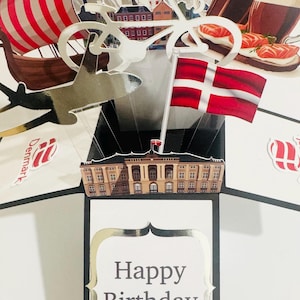 May include: A pop-up birthday card with a silver "Happy Birthday" message. The card features Danish flags, a Viking ship, and images of Danish food and architecture. The card is white with black and silver accents.