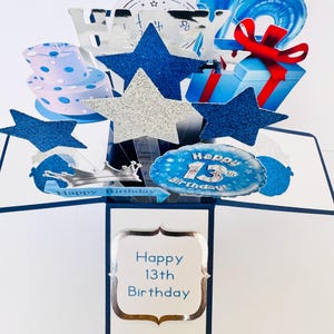 Puede incluir: Una tarjeta emergente azul y plateada con un diseño 3D que presenta un pastel, estrellas, globos y una caja de regalo. La tarjeta dice "Happy 13th Birthday".