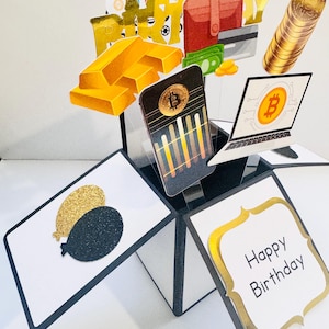 May include: A pop-up birthday card with a gold and black theme. The card features a variety of images, including gold bars, a laptop, a wallet, and a credit card. The card also includes the text "Happy Birthday".