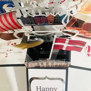 May include: A pop-up birthday card with a silver "Happy Birthday" cut-out. The card features images of a Danish flag, buildings, and a plane. The bottom of the card has the words "Happy Birthday" in a decorative frame.