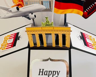 May include: A 3D pop-up greeting card featuring a German theme with a central Brandenburg Gate cutout, a German flag, a white airplane, a beer mug, an accordion, and a passport. The card displays the text Happy Birthday in metallic lettering.