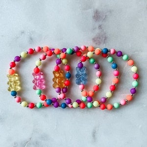 May include: Five colorful beaded bracelets with gummy bear charms. The bracelets are made of small, round beads in shades of pink, orange, yellow, green, and blue. Each bracelet has a single gummy bear charm in a different color.