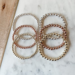 May include: Four beaded bracelets in silver, gold, and rose gold. The bracelets are stacked on top of each other.