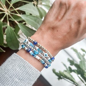 May include: A set of five bracelets with blue and white evil eye beads. The bracelets are stacked on a person's wrist.