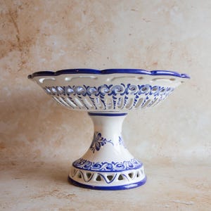 May include: A white ceramic pedestal bowl with blue floral patterns and a scalloped edge. The bowl features a pierced design and a pedestal base, ideal for displaying fruit or decorative items.